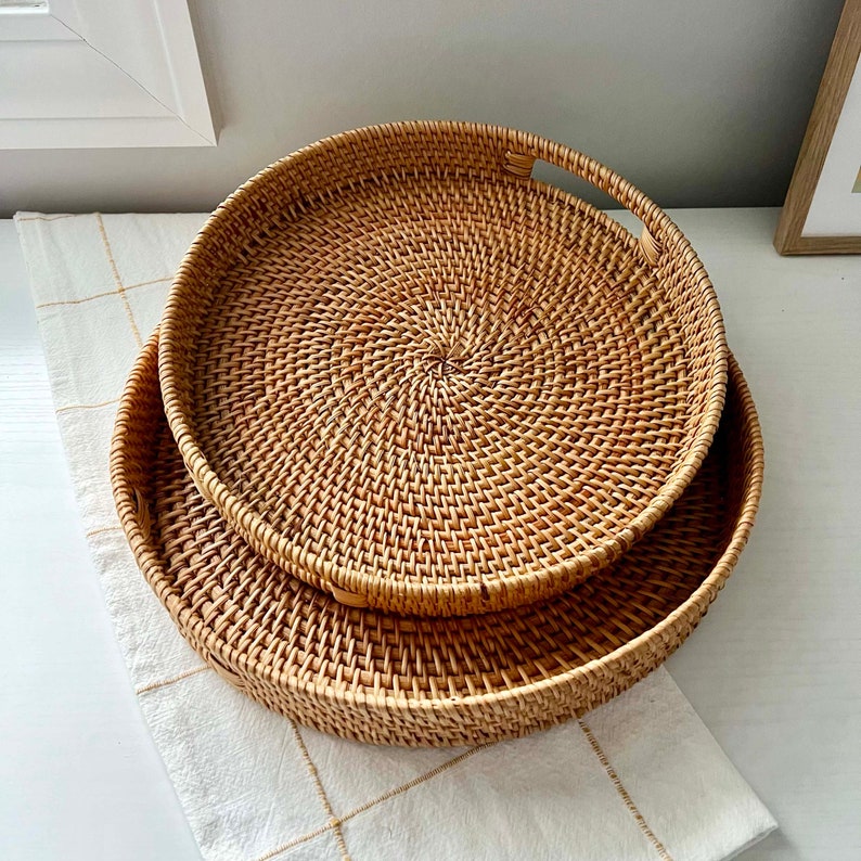 Op de afbeelding: Twee ronde, geweven rotan trays in natuurlijke tinten. De trays zijn genest, waarbij de ene iets groter is dan de andere. De trays hebben een spiraalvormig weefpatroon en zijn geplaatst op een wit oppervlak met een cr&egrave;mekleurige, geblokte doek.