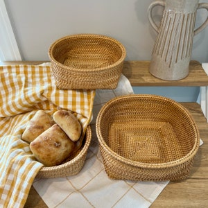 May include: Three woven rattan baskets of varying sizes. One basket holds several loaves of bread on a yellow and white checkered cloth. The other two baskets are empty. A beige pitcher sits on a wooden shelf in the background.