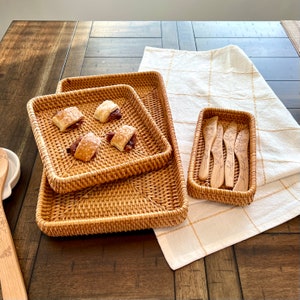 Puede incluir: Bandejas de servicio de ratán tejido de varios tamaños, apiladas sobre una mesa de madera. Las bandejas contienen pasteles. Una pequeña cesta rectangular contiene espátulas de madera con diseños florales. También hay un paño a cuadros blanco y beige.