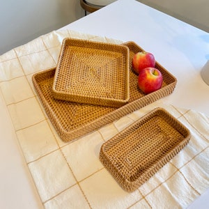 May include: A set of four woven rattan trays in varying sizes. The trays are rectangular and square, with a natural light brown color. Two red apples rest on one of the trays. The trays are arranged on a cream-colored, checkered tablecloth.