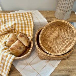 May include: Two woven rattan bread baskets, one filled with fresh bread rolls on a yellow and white checkered cloth. A second basket is stacked on top of another, with a white and gold checkered cloth underneath.