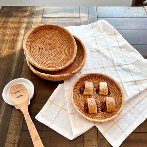 May include: Two round, woven rattan serving trays are stacked on a dark wood table. A smaller tray holds pastries. A white ceramic spoon rest and a wooden spoon with a honeycomb design are also present. A white and gold checkered cloth is draped on the table.