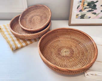 May include: A set of four round, woven rattan baskets in varying sizes. The baskets are light brown with a spiral weave pattern. One basket is on a white surface, with the other three stacked on a yellow and white checkered cloth.