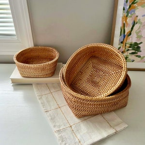 May include: Three woven rattan baskets in varying sizes. The baskets are oval-shaped and have a natural, light brown color. One basket sits on a stack of books, while the other two are stacked on a cream-colored, checkered cloth.