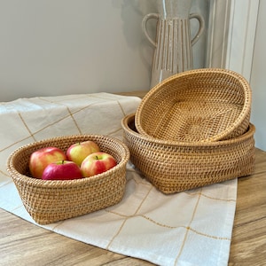 May include: Three woven rattan baskets of varying sizes are stacked on a white and gold checkered tablecloth. The largest basket contains four red and yellow apples. A beige ceramic vase is in the background.