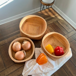May include: Three woven rattan baskets of varying sizes. The baskets contain onions and bell peppers. A white and gold patterned cloth is draped near the baskets. The baskets are on a wooden surface.
