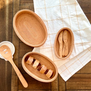 May include: An overhead shot of a wooden table with several woven rattan oval baskets. One basket holds pastries, another holds wooden knives, and a third is empty. A wooden spoon with a honeycomb design rests on a white ceramic spoon rest.