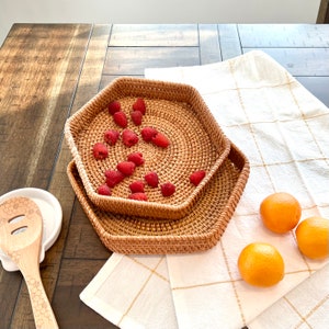Könnte beinhalten: Zwei sechseckige Tabletts aus geflochtenem Rattan, eines auf dem anderen gestapelt, mit frischen roten Himbeeren auf dem oberen Tablett. Ein Holzlöffel und Orangen sind ebenfalls zu sehen, was auf eine Küche oder einen Essbereich hindeutet.