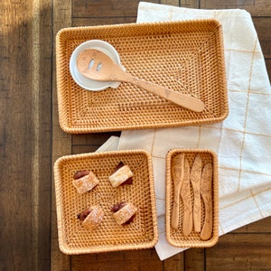 May include: A collection of woven rattan serving trays and utensils. The set includes a rectangular tray, a square tray with pastries, and a smaller rectangular tray with wooden butter knives. A wooden spoon rests on a white ceramic spoon rest.