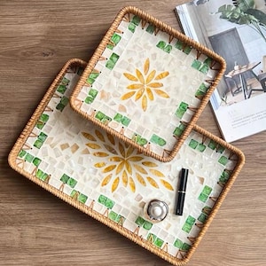 May include: Two rectangular trays with woven rattan edges and a mosaic surface featuring white shell tiles, green square accents, and a central yellow floral pattern. The larger tray holds a small silver container and a black cylindrical object.