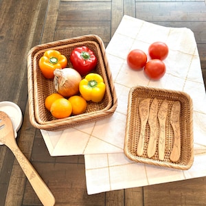 May include: A still-life image featuring woven rattan baskets filled with fresh produce. The baskets contain orange and yellow bell peppers, oranges, and an onion. Tomatoes rest on a white and gold checkered cloth, and a small basket holds wooden spreaders.