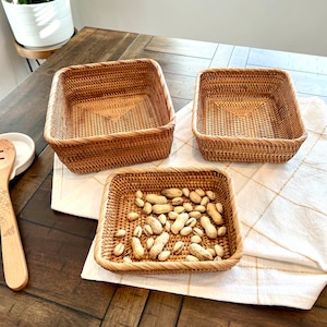 May include: Three woven rattan baskets of varying sizes. One basket is filled with peanuts and pistachios. The baskets are square-shaped with a natural brown color. They are placed on a white cloth with a gold pattern.