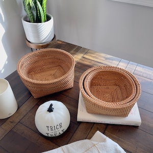 May include: Three woven rattan baskets of varying sizes, nested together, sit on a wooden table. A white pumpkin with "Trick or Treat" text is nearby. A potted plant and a book are also on the table.