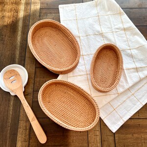 May include: A collection of woven rattan oval trays in varying sizes, alongside a wooden spoon with a bee design and a white ceramic spoon rest. A white and gold checkered cloth is also present.