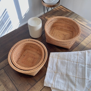 Könnte beinhalten: Drei gewebte Rattankörbe unterschiedlicher Größe sind auf einem dunklen Holztisch ineinander verschachtelt. Eine weiße Kerze steht daneben, und ein weißes und goldfarbenes kariertes Tuch ist auf dem Tisch drapiert. Die Körbe sind leer und einsatzbereit.