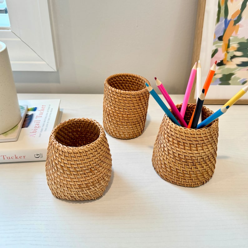 Pu&ograve; includere: Tre portapenne in rattan intrecciato di varie dimensioni. Un portapenne contiene diverse matite colorate. I portapenne sono di colore marrone chiaro e hanno un design a trama di vimini. I portapenne sono su una superficie bianca.