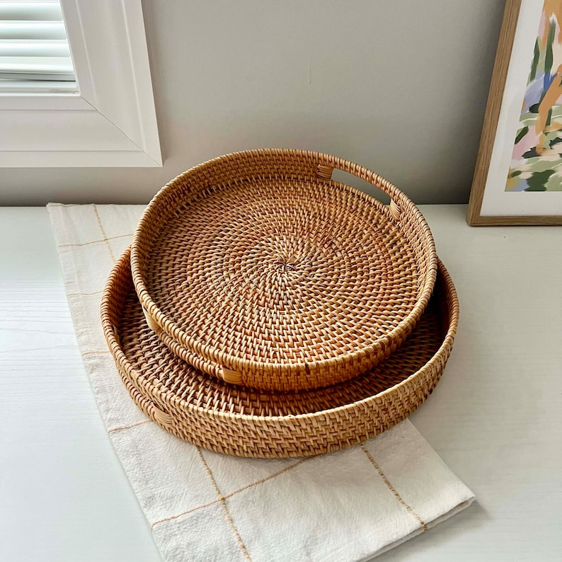 Op de afbeelding: Twee ronde, geweven rotan trays in verschillende maten. De trays zijn lichtbruin en hebben een natuurlijke, getextureerde uitstraling. De grotere tray heeft handvatten. Ze zijn geplaatst op een cr&egrave;mekleurige, geruite stof.