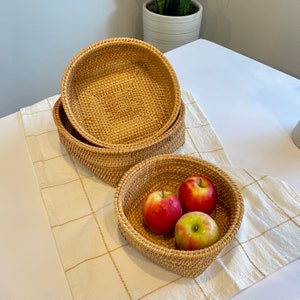 May include: Three woven rattan baskets, two stacked and one holding three red and green apples. The baskets are round with a woven pattern and sit on a cream-coloured tablecloth with a grid pattern.