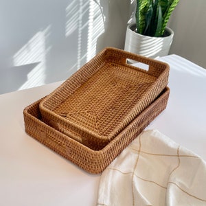 May include: A set of two rectangular woven rattan trays with a natural light brown finish. Each tray features a structured grid pattern and integrated cutout handles on the shorter sides, with one tray nested inside the other for storage.
