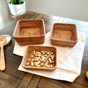 May include: Three square woven rattan baskets of different sizes. The largest and medium baskets are empty, while the smallest contains peanuts. The baskets sit on a white cloth with a gold grid pattern.