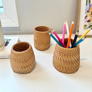 Pu&ograve; includere: Tre portapenne in rattan intrecciato di diverse dimensioni sono su una superficie bianca. Il portapenne pi&ugrave; grande contiene diverse matite colorate. I portapenne sono di colore beige naturale e hanno un design a trama di vimini. Ideali per l'organizzazione della scrivania.