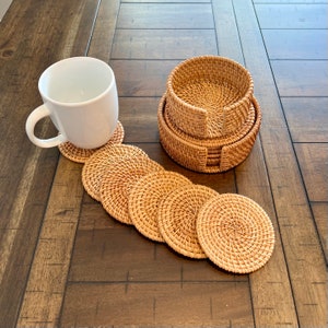 May include: A set of woven rattan coasters and a matching holder. The coasters are round and have a natural, light brown color. A white mug sits on one of the coasters. The coasters are arranged on a dark wooden surface.