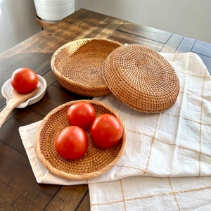 May include: A collection of woven rattan baskets and a round woven lid. Three red tomatoes rest in a small basket. A white spoon rest with a wooden spoon and a single tomato are also present. The items are on a wooden table with a white and gold checkered cloth.