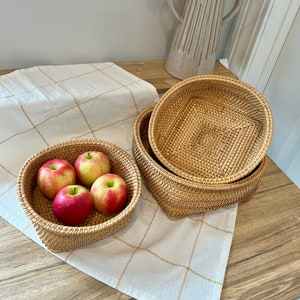 May include: Two woven rattan baskets, one round and one square, are stacked on a white and gold checkered tablecloth. The round basket contains four red and yellow apples. A beige ceramic pitcher is in the background.