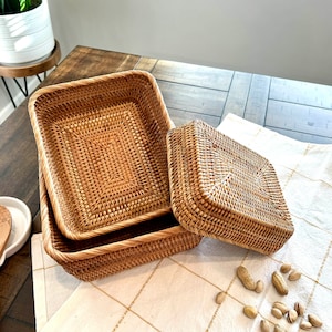 May include: Three woven rattan baskets in varying sizes. The rectangular baskets are stacked, with one open and the other nested inside. A square basket with a lid sits beside them. The baskets are a warm, natural brown color.
