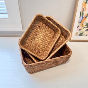 Könnte beinhalten: Drei rechteckige Körbe aus geflochtenem Rattan in verschiedenen Größen sind ineinander verschachtelt. Die Körbe haben eine natürliche, hellbraune Farbe und eine strukturierte Oberfläche. Die Kanten sind mit einem dickeren geflochtenen Rand verstärkt. Die Körbe sind auf einer weißen Oberfläche angeordnet.