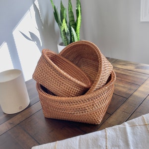 Puede incluir: Tres cestas de ratán tejido de diferentes tamaños están apiladas sobre una mesa de madera oscura. Las cestas son de color marrón claro natural y tienen un tejido abierto texturizado. Una vela blanca y una planta de interior están en el fondo.