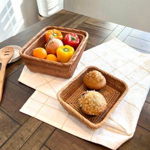 May include: Two woven rattan baskets on a wooden table. One basket contains oranges, an onion, and red, yellow, and orange bell peppers. The other basket holds two muffins. The baskets sit on a white and gold checkered tablecloth.