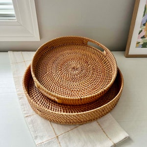 Può includere: Due vassoi rotondi in rattan intrecciato di diverse dimensioni. I vassoi marrone chiaro hanno manici. Sono posizionati su una superficie bianca con un panno a scacchi color crema. Un'opera d'arte incorniciata è visibile sullo sfondo.