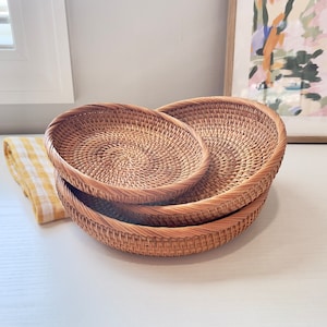 May include: Three round, woven rattan baskets of varying sizes are stacked on a white surface. The baskets have a natural, light brown color and a textured surface. A yellow and white checkered cloth is visible on the left side of the image.