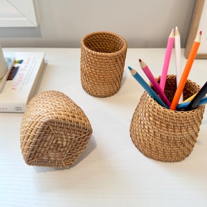 Pu&ograve; includere: Tre organizzatori da scrivania in rattan intrecciato. Uno &egrave; un cestino rettangolare, un altro &egrave; un portapenne cilindrico, e il terzo &egrave; un portapenne con matite colorate all'interno. Gli organizzatori sono marrone chiaro e poggiano su una superficie bianca.