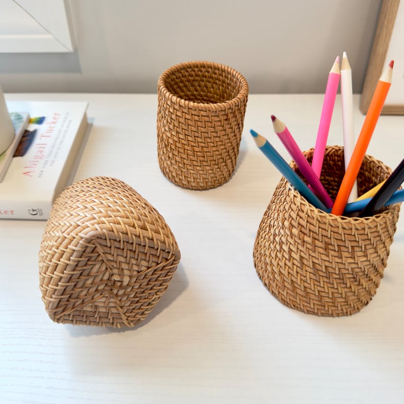 Pu&ograve; includere: Tre organizzatori da scrivania in rattan intrecciato. Uno &egrave; un cestino rettangolare, un altro &egrave; un portapenne cilindrico, e il terzo &egrave; un portapenne con matite colorate all'interno. Gli organizzatori sono marrone chiaro e poggiano su una superficie bianca.