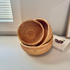 May include: Three round, woven rattan baskets of varying sizes are nested together. The baskets have a natural, light brown color and a textured surface. The rims of the baskets are slightly raised and have a braided design. A book is visible in the background.