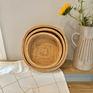 May include: Three round, woven rattan baskets of different sizes nested together. The baskets are light brown with a square pattern in the centre. A white and tan checked cloth is beneath the baskets. A vase with yellow flowers is in the background.