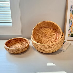 May include: Two round, woven rattan baskets of different sizes. The larger basket is nested inside the smaller one. The baskets are light brown and have a natural, textured appearance. They are placed on a white surface.