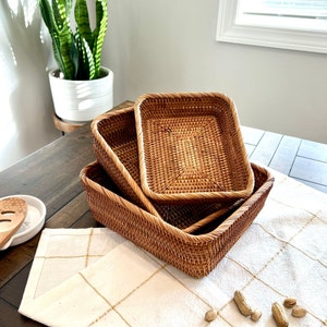 May include: Three rectangular woven baskets in different sizes are stacked on a table. The baskets are a warm brown colour and have a textured surface. A white and gold checked tablecloth is beneath the baskets. A potted plant is in the background.