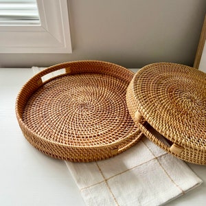 Könnte beinhalten: Zwei runde Tabletts aus geflochtenem Rattan. Ein Tablett ist offen, mit Griffen auf beiden Seiten. Das andere Tablett hat einen Deckel. Beide Tabletts sind hellbraun und liegen auf einer weißen Oberfläche mit einem cremefarbenen, karierten Stoff.