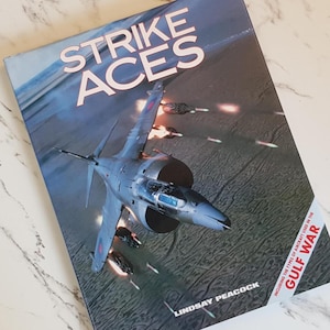 May include: A book titled "Strike Aces" with a jet fighter on the cover, set against a backdrop of explosions. The cover is predominantly blue and grey, with the title in white. Author's name, Lindsay Peacock, is visible.