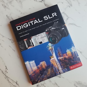 May include: A book titled "Mastering Your Digital SLR" by Chris Weston. The cover shows a Nikon digital camera and a cityscape with a blue sky. The book is published by RotoVision.