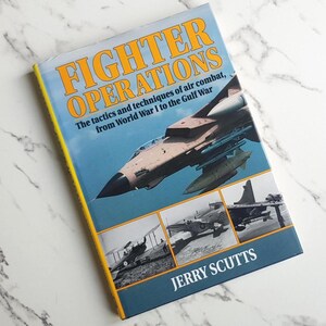 May include: A book titled "FIGHTER OPERATIONS" with a blue cover showing a fighter jet and other aircraft. The title is in orange, with the author's name, Jerry Scutts, at the bottom. The book covers air combat tactics from World War I to the Gulf War.