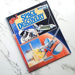 May include: A hardback book titled Space Discovery with the Eye Openers logo. The cover features illustrations of an astronaut, a space shuttle, a rocket launching, and a satellite against a backdrop of space and a planet, with the publisher name Macdonald.