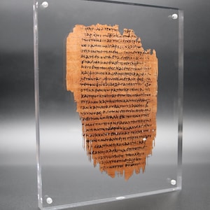 May include: A framed ancient papyrus fragment with visible text. The papyrus is a light brown color, and the text is written in black ink. The frame is clear acrylic, held together with four silver screws. The papyrus is displayed against a gray background.