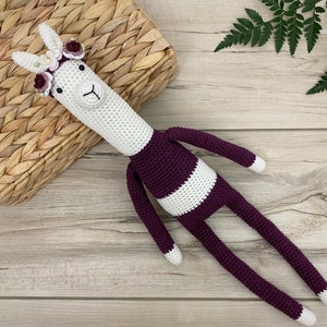 May include: A white crocheted llama with a purple and white striped body and a flower crown. The llama is lying on a wooden surface.