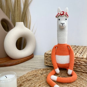 May include: A crocheted llama toy with a white body and orange legs and a flower crown. The llama is sitting on a woven basket.