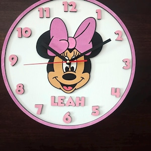 May include: A round wall clock with a white face and a pink border. The clock features a central illustration of a cartoon mouse with a pink bow, black ears, and a smiling face. The name LEAH is printed in pink below the mouse. Black hour and minute hands and a red second hand are present, with raised pink numbers from 1 to 12.