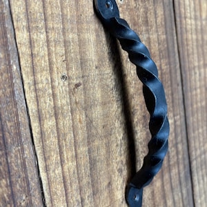 May include: Black, twisted metal handle with a decorative design. The handle is attached to a wooden surface with a natural wood grain. The handle is a decorative element for furniture or doors.